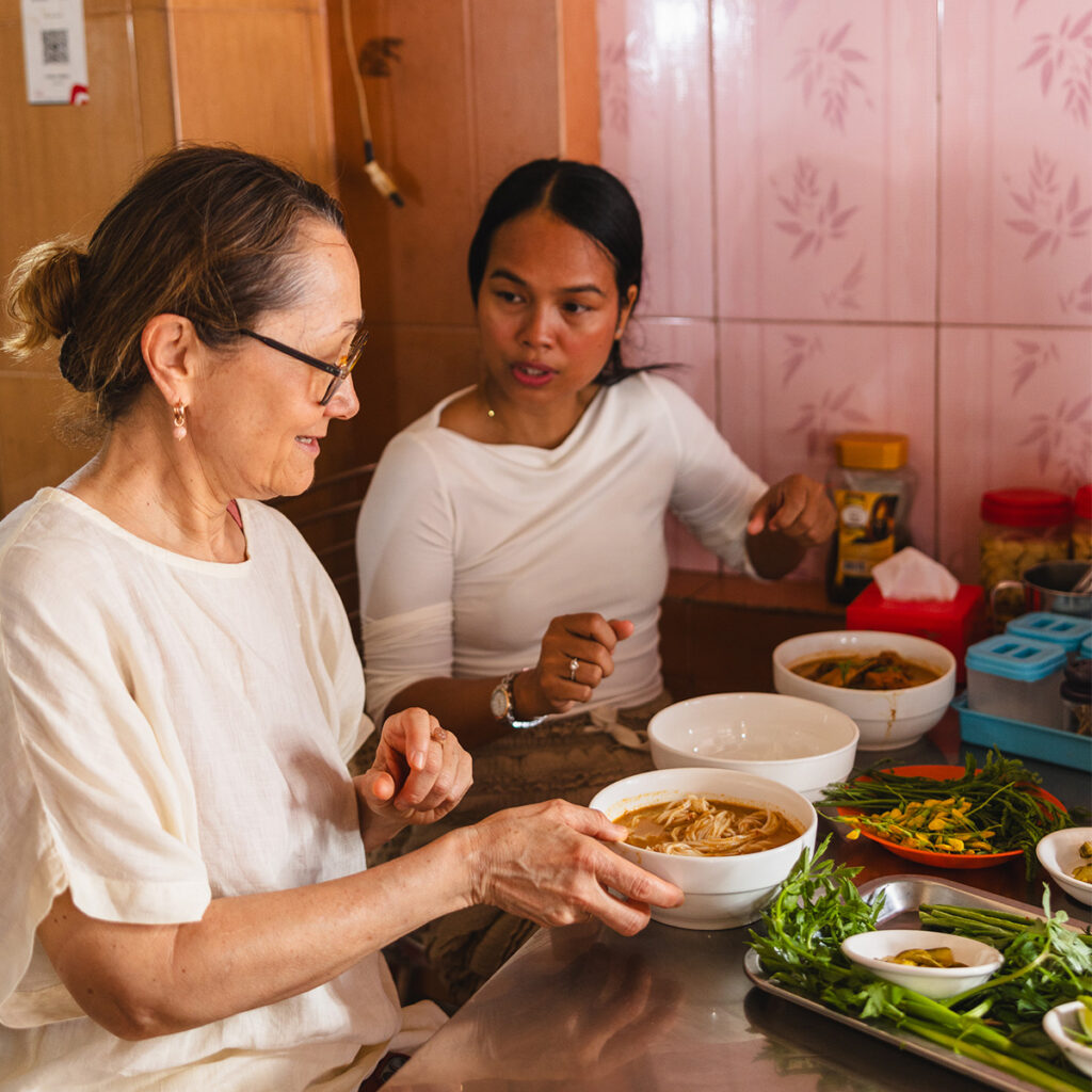 Two women working in a kitchen making noodle soup, with bowls, herbs, and prep ingredients on the counter.
