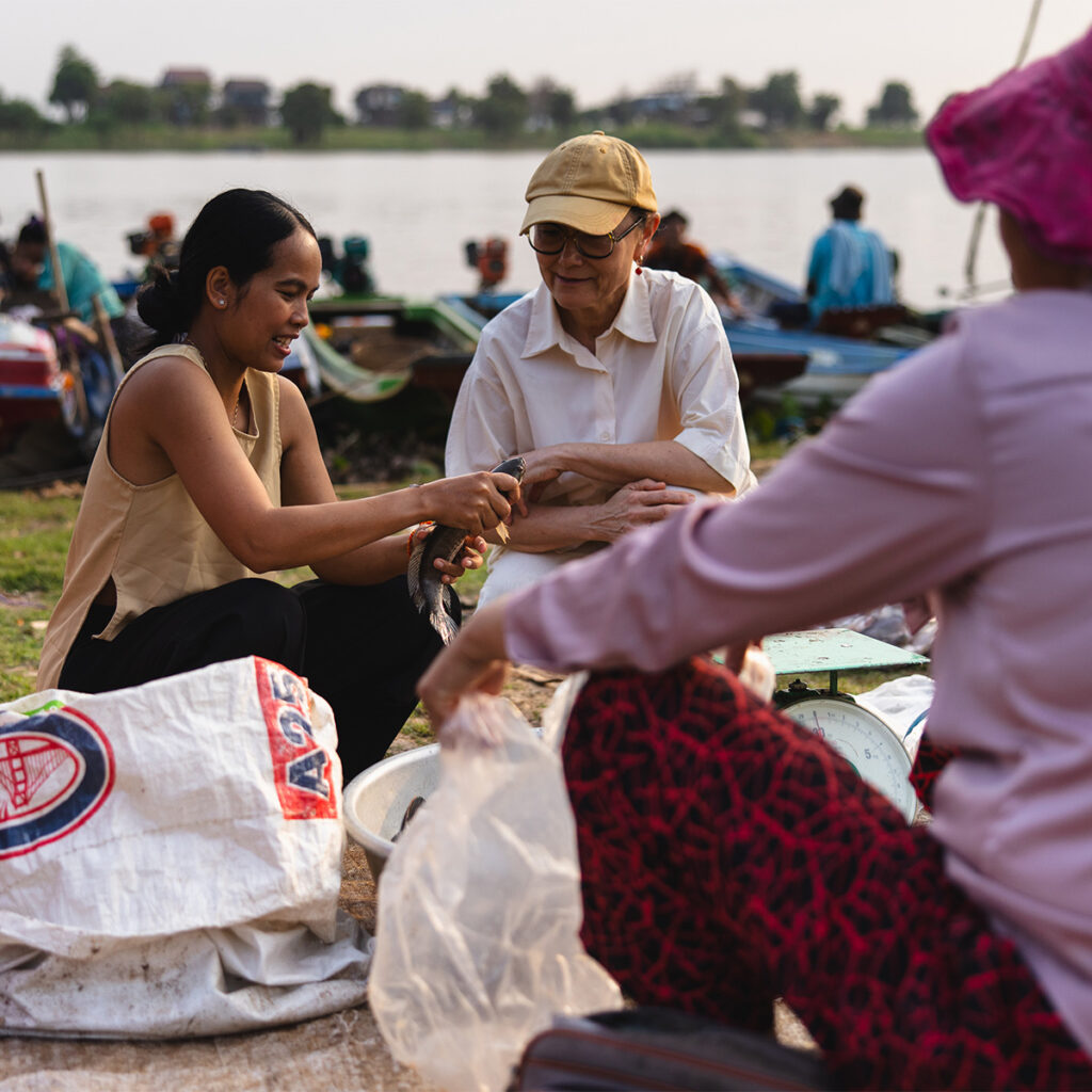 Two women sit by a riverside market, weighing and handling a fish with bags and a scale nearby.