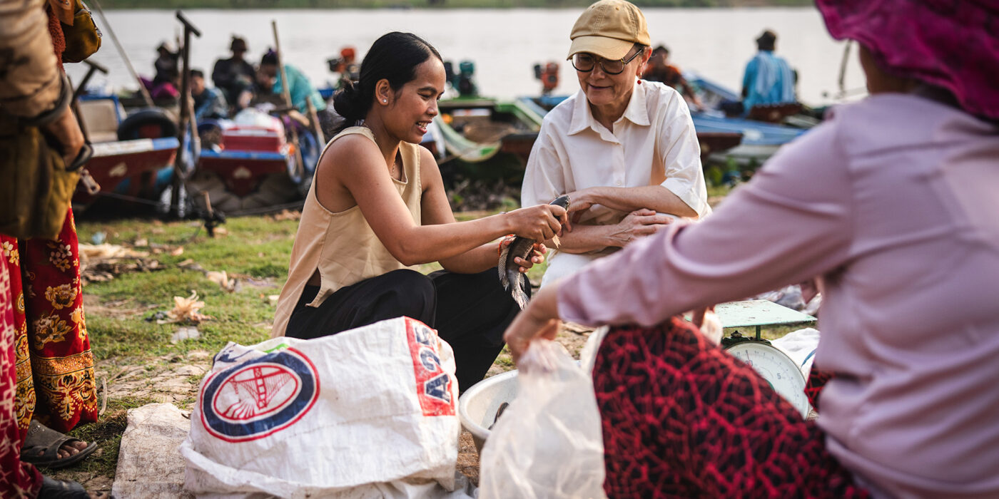 Two women exchange a fish on a riverbank market, with sacks, scales, and boats in the background.