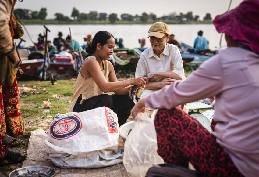 Two women exchange a fish on a riverbank market, with sacks, scales, and boats in the background.