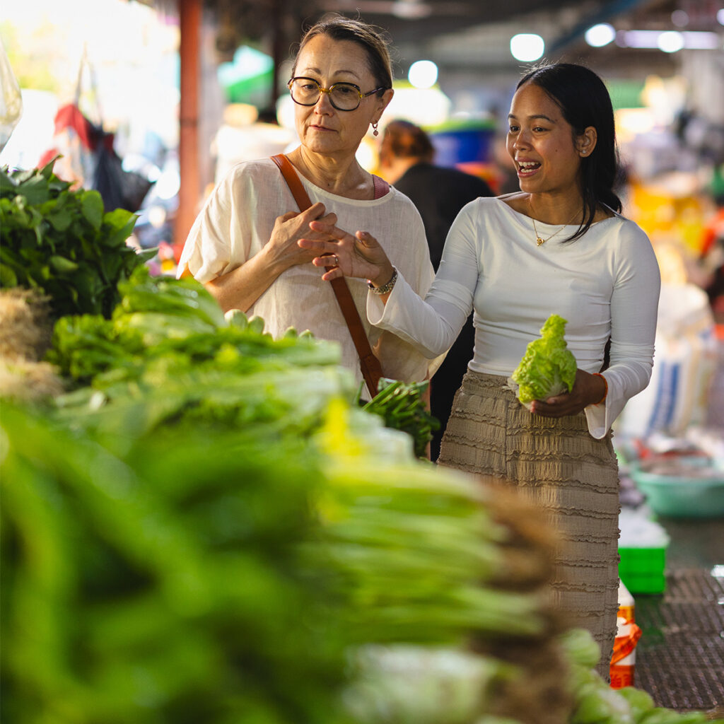 Two women at a market stall with fresh greens; one holds lettuce while the other gestures and smiles.