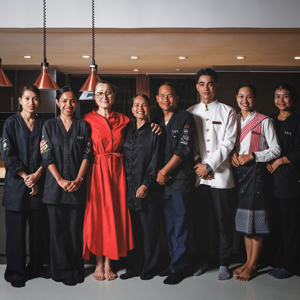 Eight people stand together in a modern kitchen studio, some in black uniforms, one in a red dress, smiling at the camera.