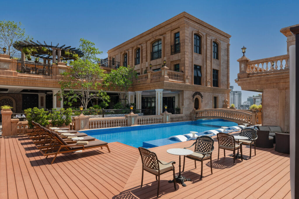 Luxurious outdoor hotel pool area with wooden deck, lounge chairs, and a beige stone building in the background under a clear blue sky.