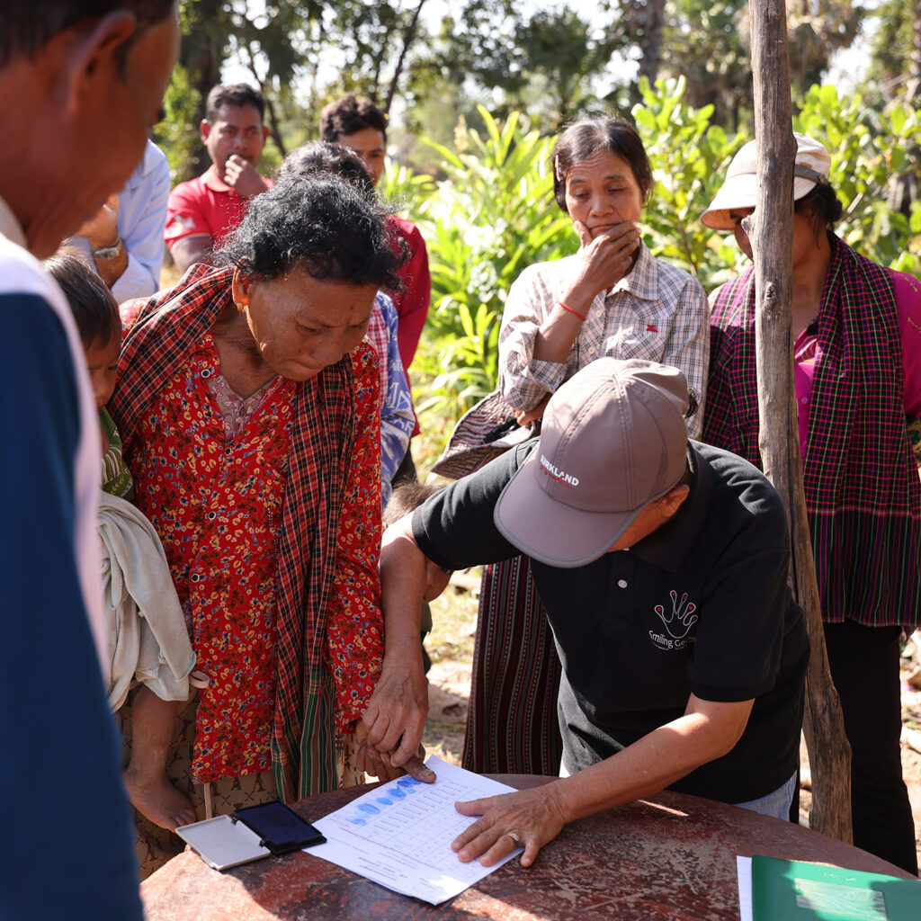 A group of villagers gathers around a weathered table outdoors, as a man in a black polo signs papers while an older woman in red floral clothing watches nearby.