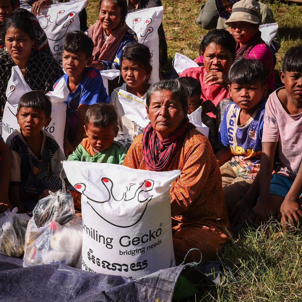 Group of people and children sit on the ground outdoors with large donation bags labeled 'Smiling Gecko'.