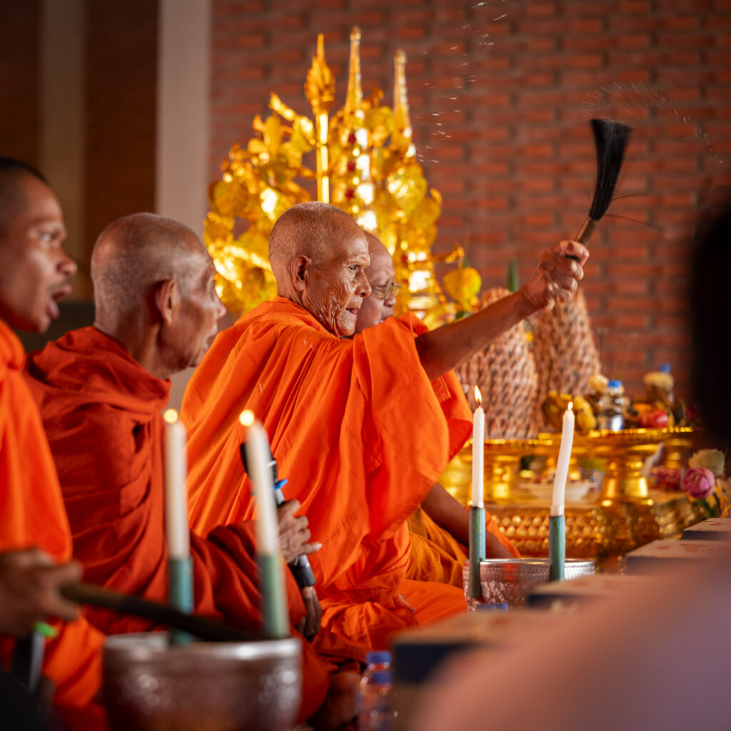 An elderly Buddhist monk in orange robes raises a ceremonial whisk during a ritual at a gold-altar with lit candles nearby.