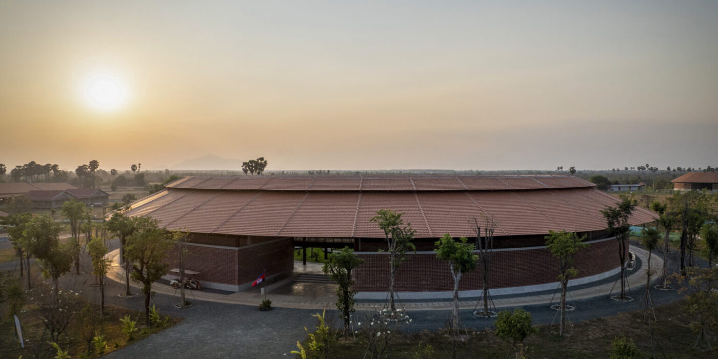 DJI_0297-1536x1229 - Smiling Gecko Circular brick building with a red tiled roof at sunset, trees lining a curved driveway in a campus-like setting