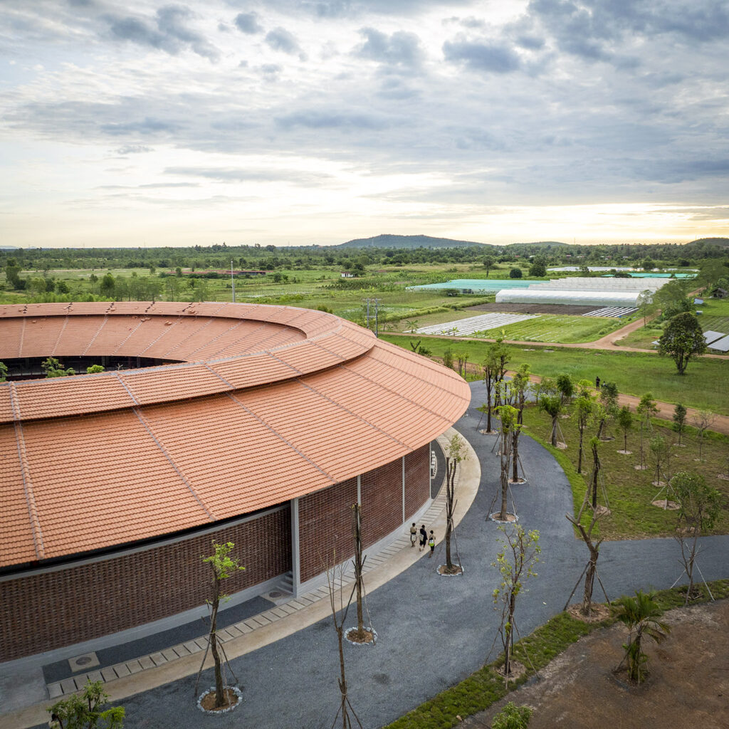 Aerial view of a circular brick building with a red tiled roof, surrounding walkway, and green fields beyond.