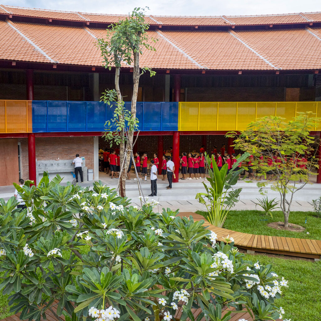 Group of students in red shirts lined up in a school courtyard, with a red-roofed building and colorful blue-yellow railing in the background.