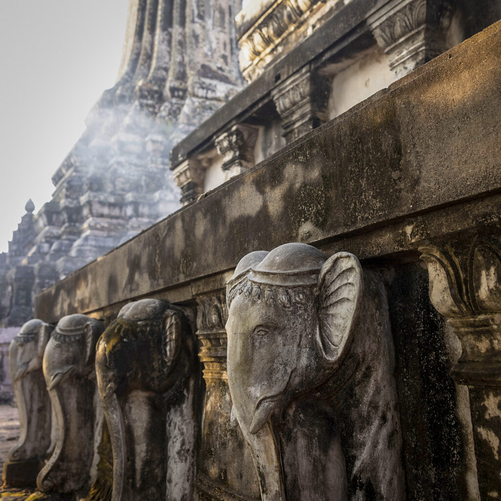 Row of weathered stone statues with winged heads along a low balustrade at an ancient temple complex, with a stone tower in the background