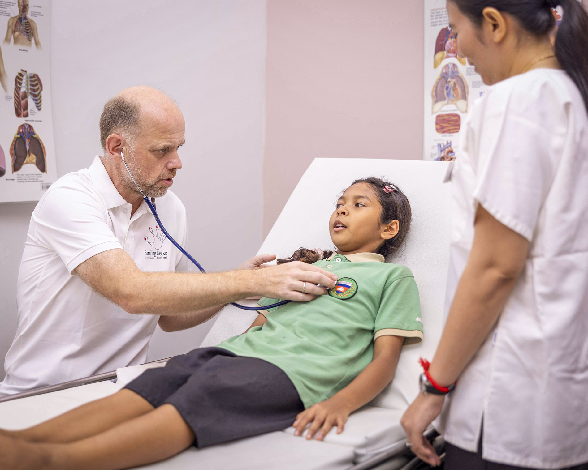 Doctor uses a stethoscope on a lying young girl during a medical checkup, with a nurse nearby in the room (clinical setting).