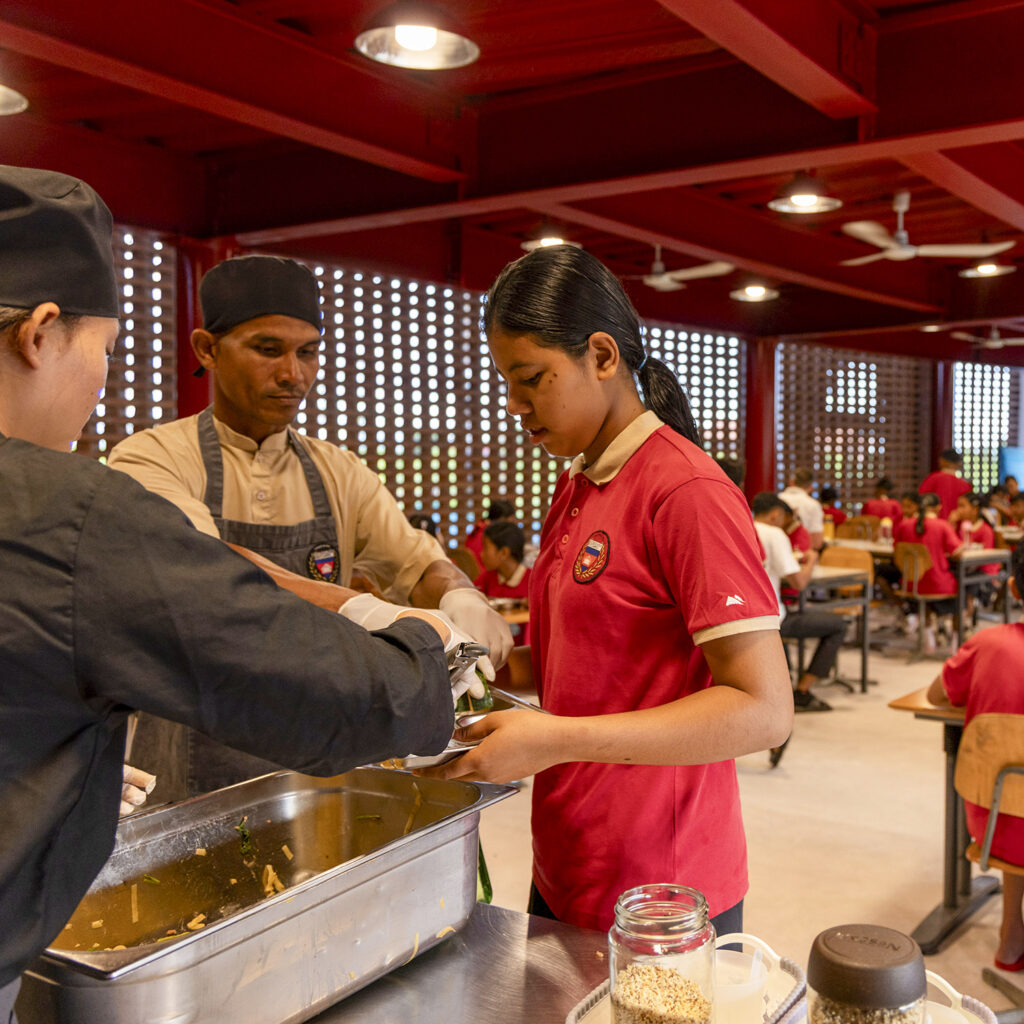 Girl in a red polo assists with food service at a cafeteria counter as staff help nearby.