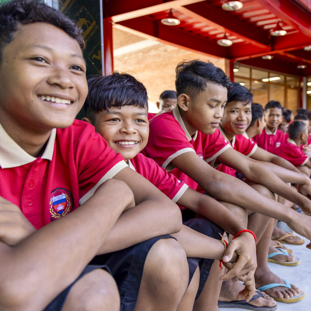 Group of smiling students in red polo shirts sitting in a row outdoors, wearing matching uniforms at a school.