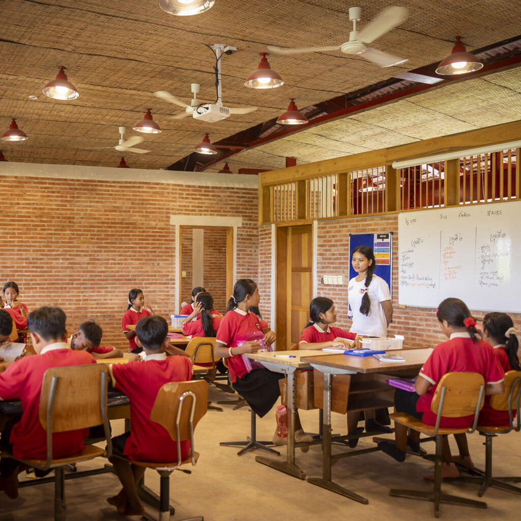 Teacher wearing white stands at a whiteboard as students in red uniforms sit at tables in a brick classroom.