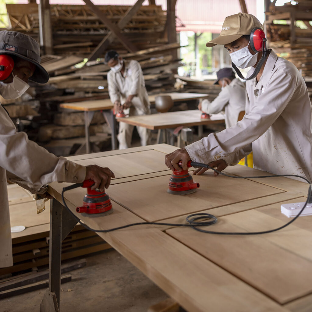 Workers in a woodshop sanding large plywood boards using red handheld sanders, wearing masks and ear protection.