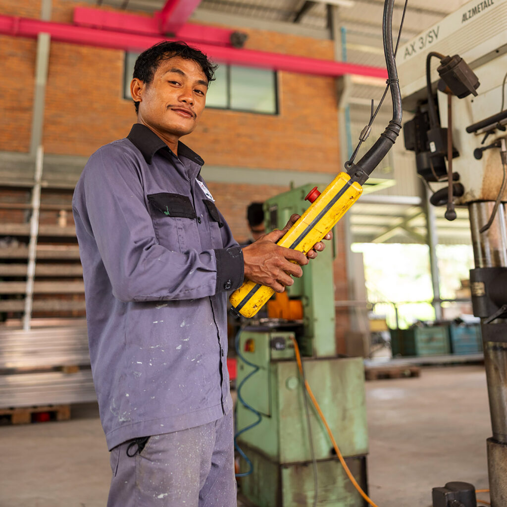 Male technician in a gray work jumpsuit holds a yellow control pendant in an industrial workshop, smiling at the camera.