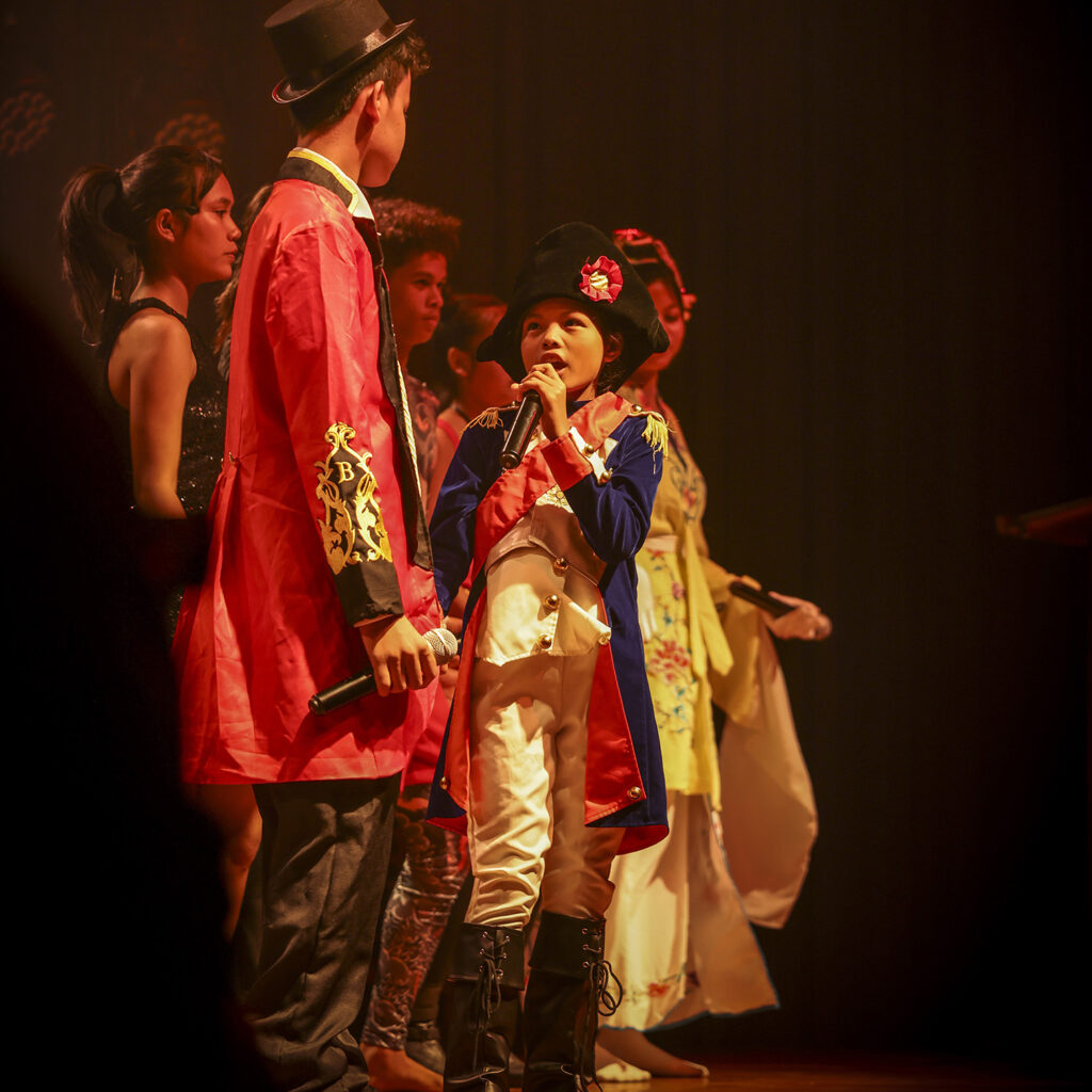 Children perform on stage in colorful costumes; a girl in a blue-red coat sings into a microphone while others wait their turn behind her.