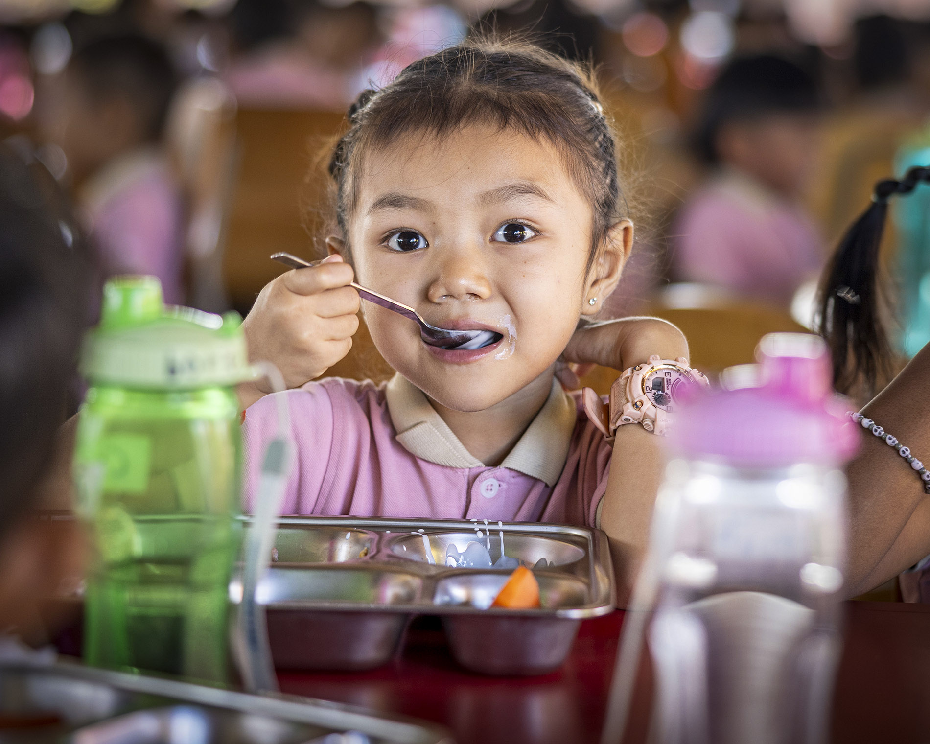 Young girl in a pink shirt eating with a spoon at a cafeteria table, looking at the camera, with a lunch tray in front.