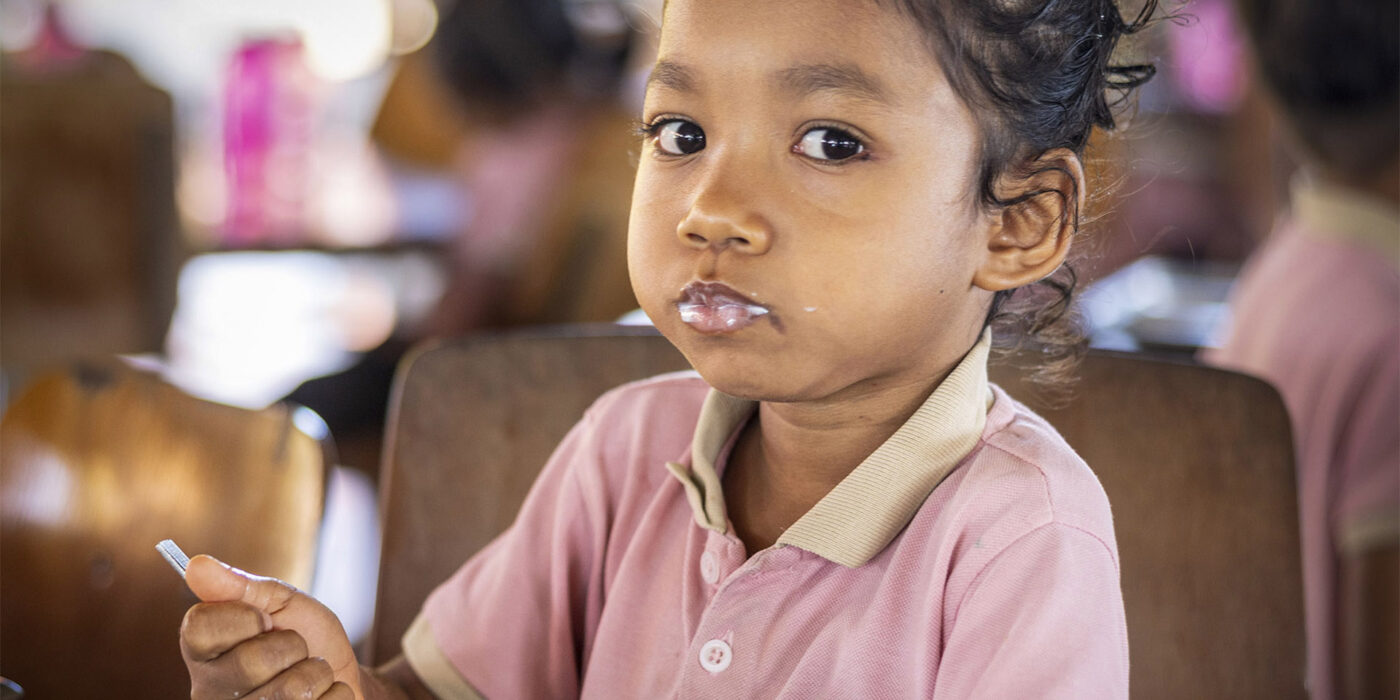 Young girl in a pink school polo sits at a table, holding a spoon with a snack, looking at the camera with milk on her lower lip.