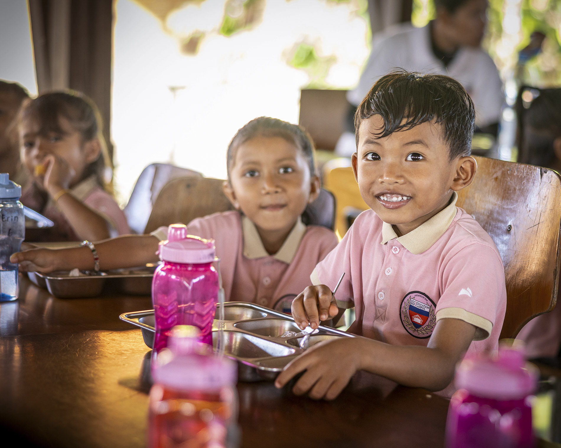 Smiling boy in a pink polo sits at a lunch table with a tray and a pink water bottle nearby.