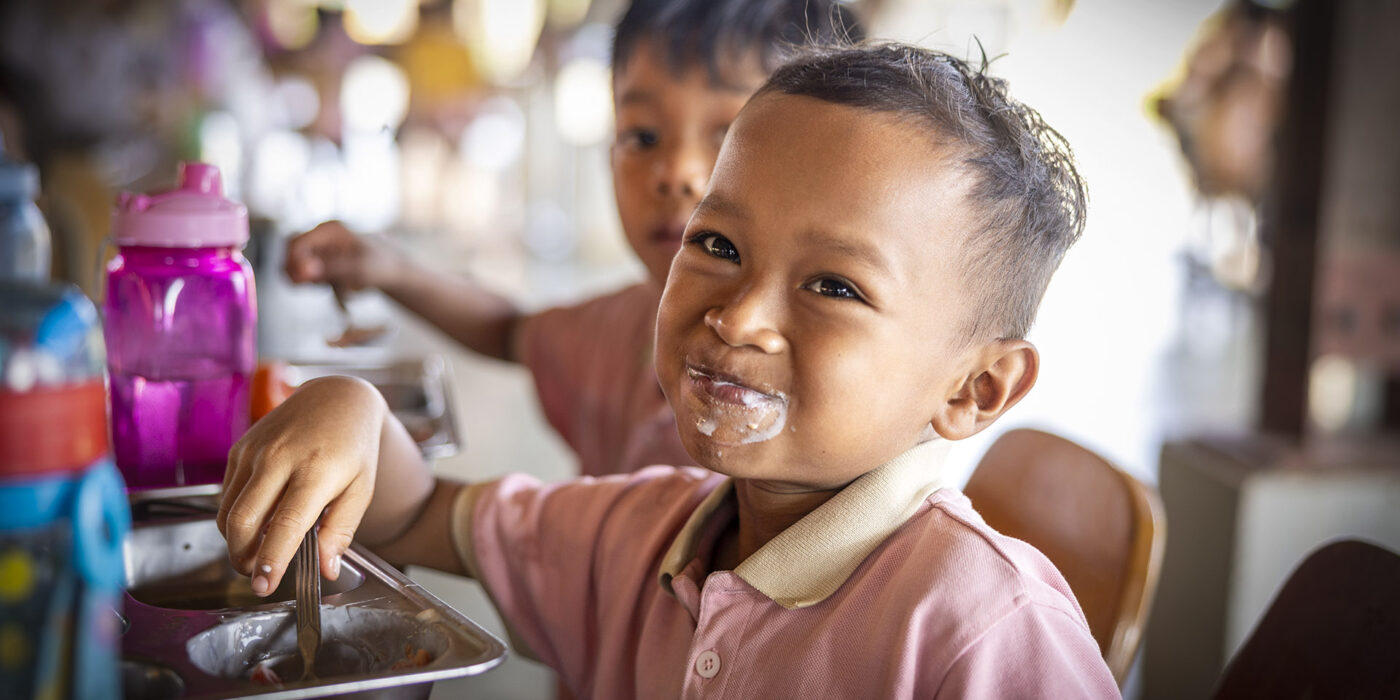 Smiling child in a pink shirt eats from a metal cafeteria tray with a spoon, food smeared around the mouth.