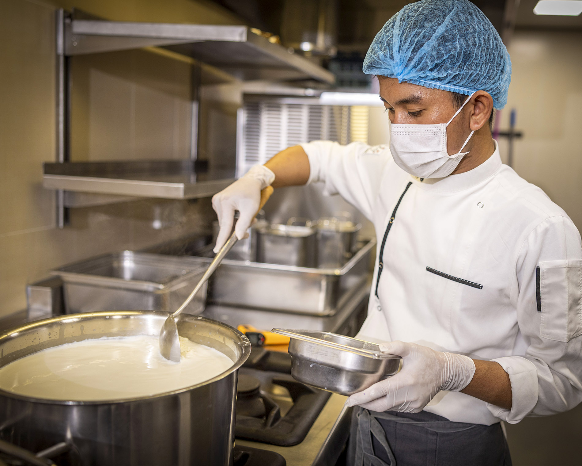 Chef wearing a blue hairnet, white mask, and gloves, ladling soup from a large stainless steel pot in a commercial kitchen