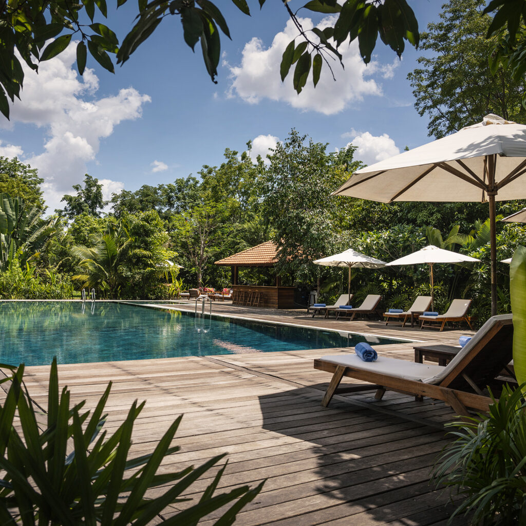 Outdoor pool area on a wooden deck with white umbrellas, lounge chairs, and lush tropical plants surrounding it.