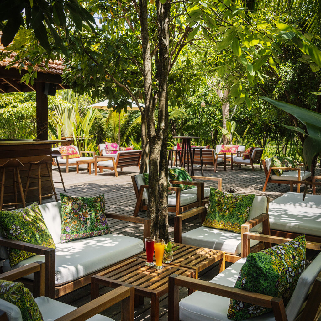 Outdoor lounge area with wooden couches and green patterned cushions under a tree, colorful drinks on a table in the foreground.