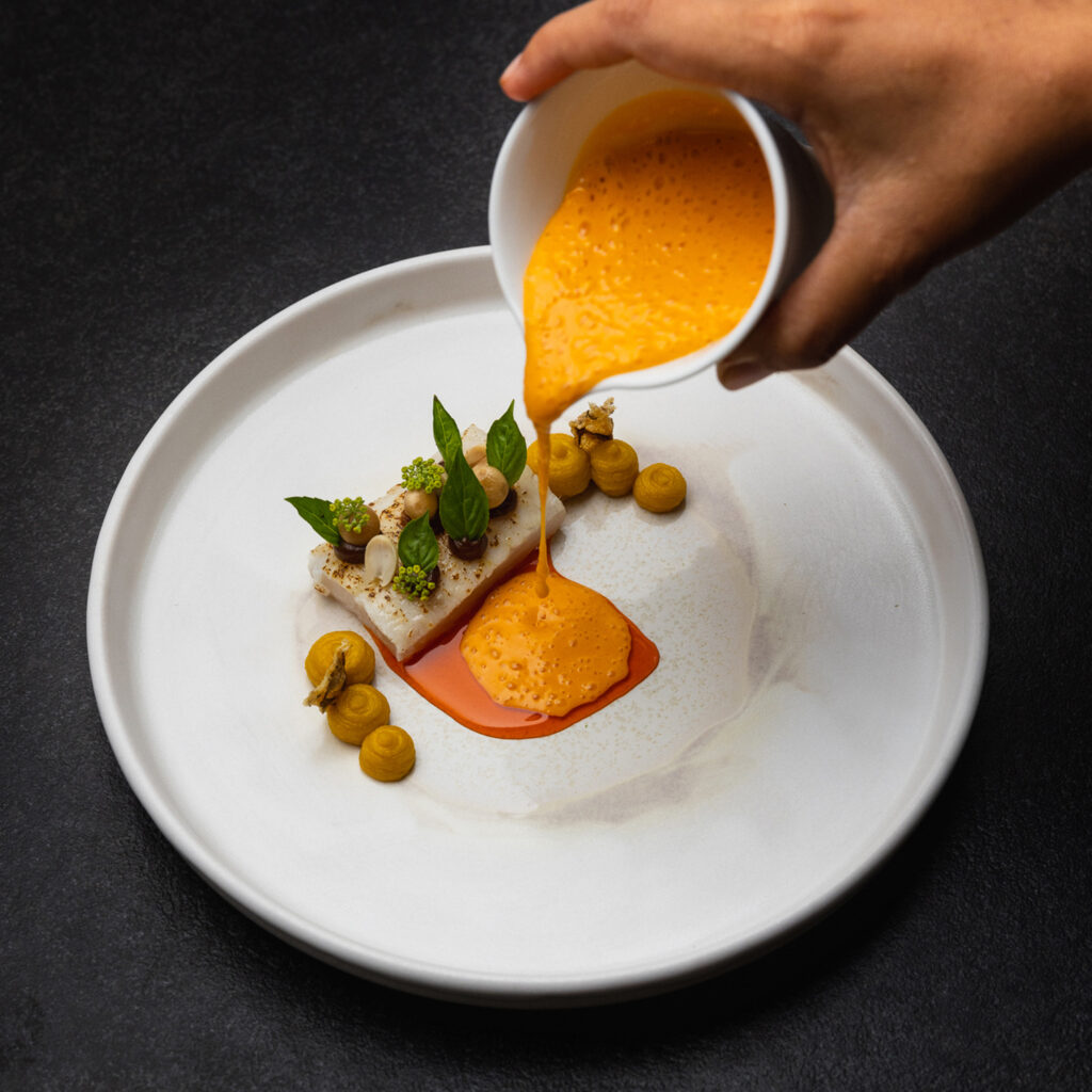 Chef pours bright orange sauce over a plated fish dish garnished with herbs and orange dollops on a white plate.