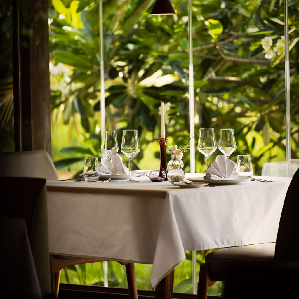 Elegant dining table set for a meal with folded white napkins, multiple wine glasses, and a tall candle centerpiece against a lush green garden visible through large glass walls.