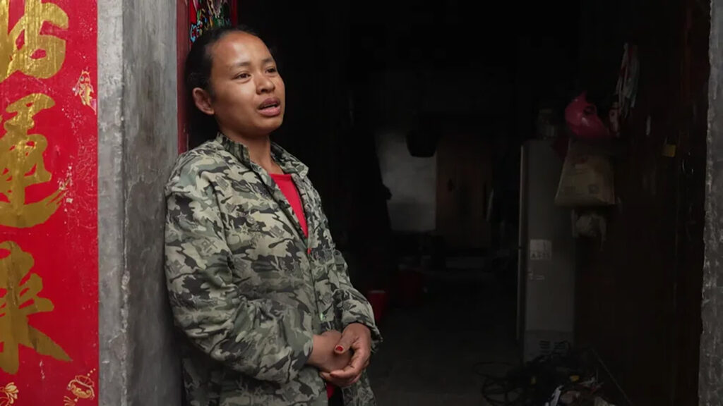 Woman in camo jacket standing in a doorway, hands clasped, looking thoughtful, with a red Chinese banner on the left and a dimly lit interior behind him/her.
