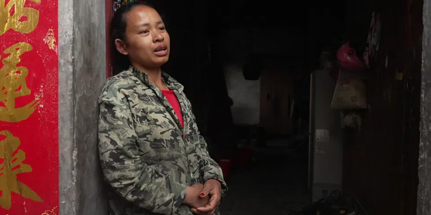 SRF - Smiling Gecko Woman in camo jacket standing in a doorway, hands clasped, looking thoughtful, with a red Chinese banner on the left and a dimly lit interior behind him/her.