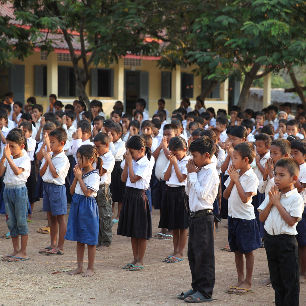 Group of uniformed children standing in rows outdoors, hands pressed together in prayer, in a schoolyard with a building behind them.
