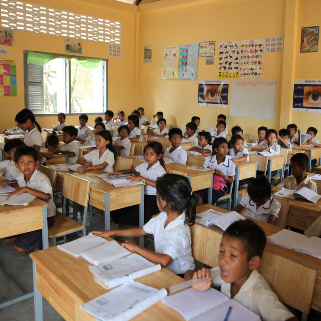 Classroom full of primary students in white uniforms at wooden desks, studying with notebooks and papers in front of them.