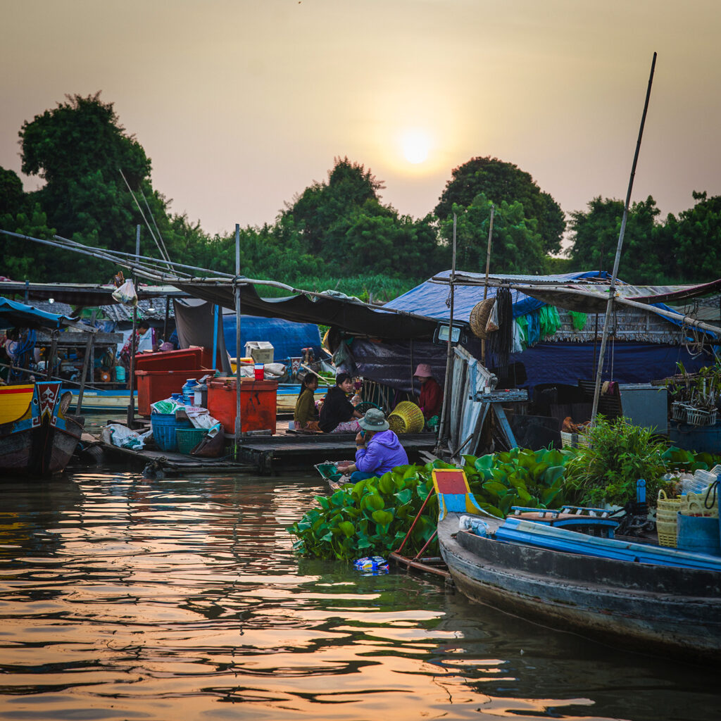 Sunset over a busy floating market with colorful boats and makeshift stalls along the riverbank.